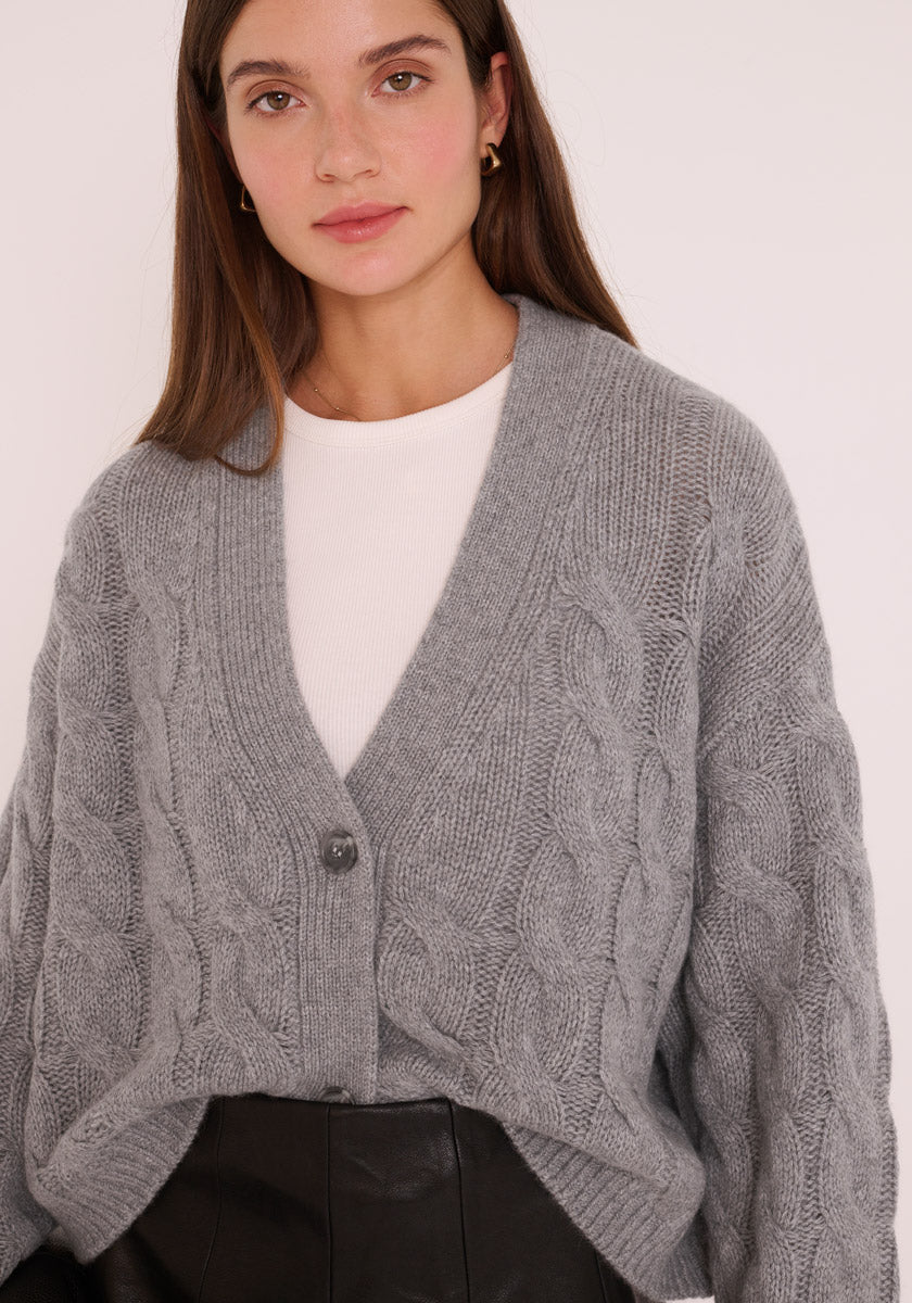 Eine Frau mit langen braunen Haaren trägt eine hellgraue Kaschmir Strickjacke von Princess Goes Hollywood mit großen Knöpfen über einem weißen Oberteil und einer schwarzen Lederhose mit hoher Taille, die sich sanft von einem hellen Hintergrund abhebt.