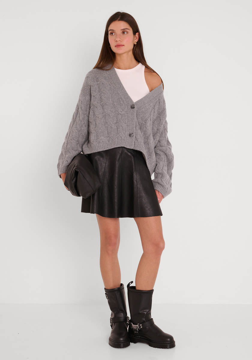 Eine Frau posiert vor einer weißen Wand in einer Princess Goes Hollywood Kaschmir Strickjacke, die über eine Schulter drapiert ist, einem weißen Top, einem schwarzen ausgestellten Lederrock, klobigen Motorradstiefeln und einer schwarzen Clutch; ihr glattes braunes Haar ist offen.