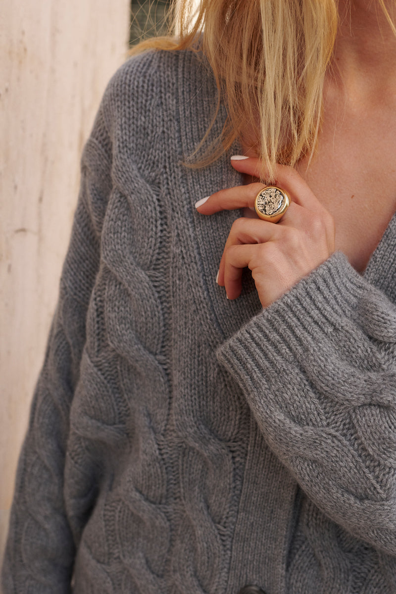 Eine Frau trägt einen dicken, grauen Princess Goes Hollywood Zopfmuster-Pullover mit tiefem V-Ausschnitt. Sie hält eine Strähne ihres blonden Haares in der linken Hand und trägt einen großen Goldring mit einem dekorativen, runden Muster. Nur ihr Oberkörper und ihr Hals sind zu sehen.