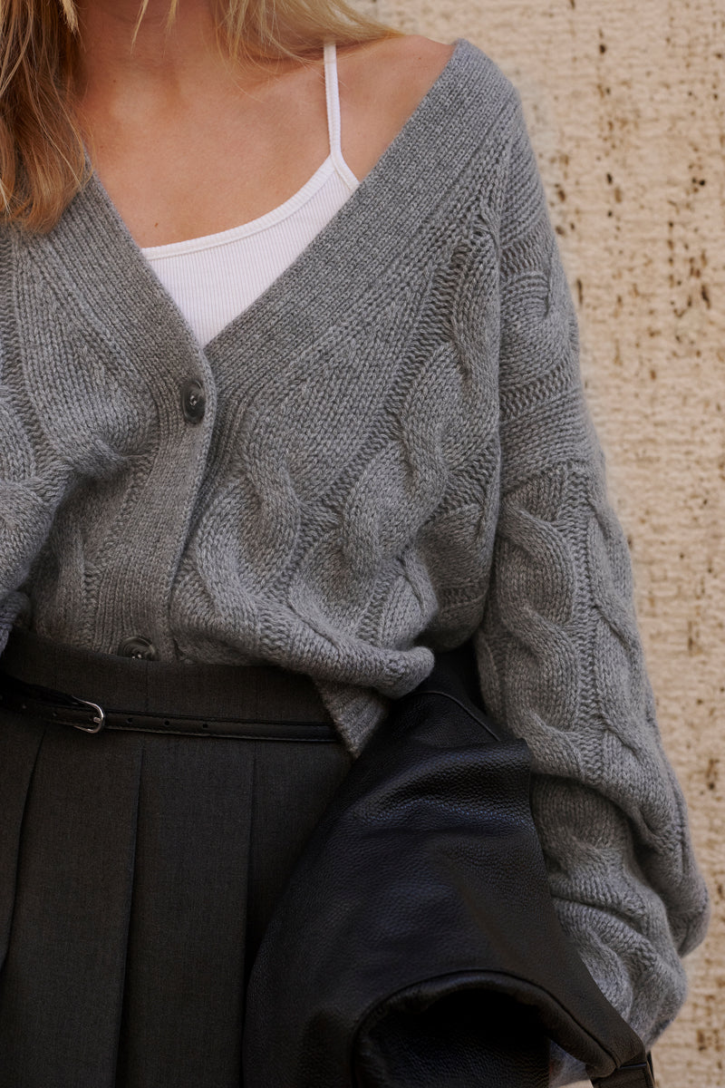 Eine Frau trägt eine klobige, hellgraue Princess Goes Hollywood Kaschmir Zopfmuster-Strickjacke mit großen Knöpfen, die über einer Schulter über ein weißes Rippentank getragen wird. Sie trägt eine schwarze strukturierte Jacke und kombiniert sie mit einer hochgezogenen Bundfaltenhose.