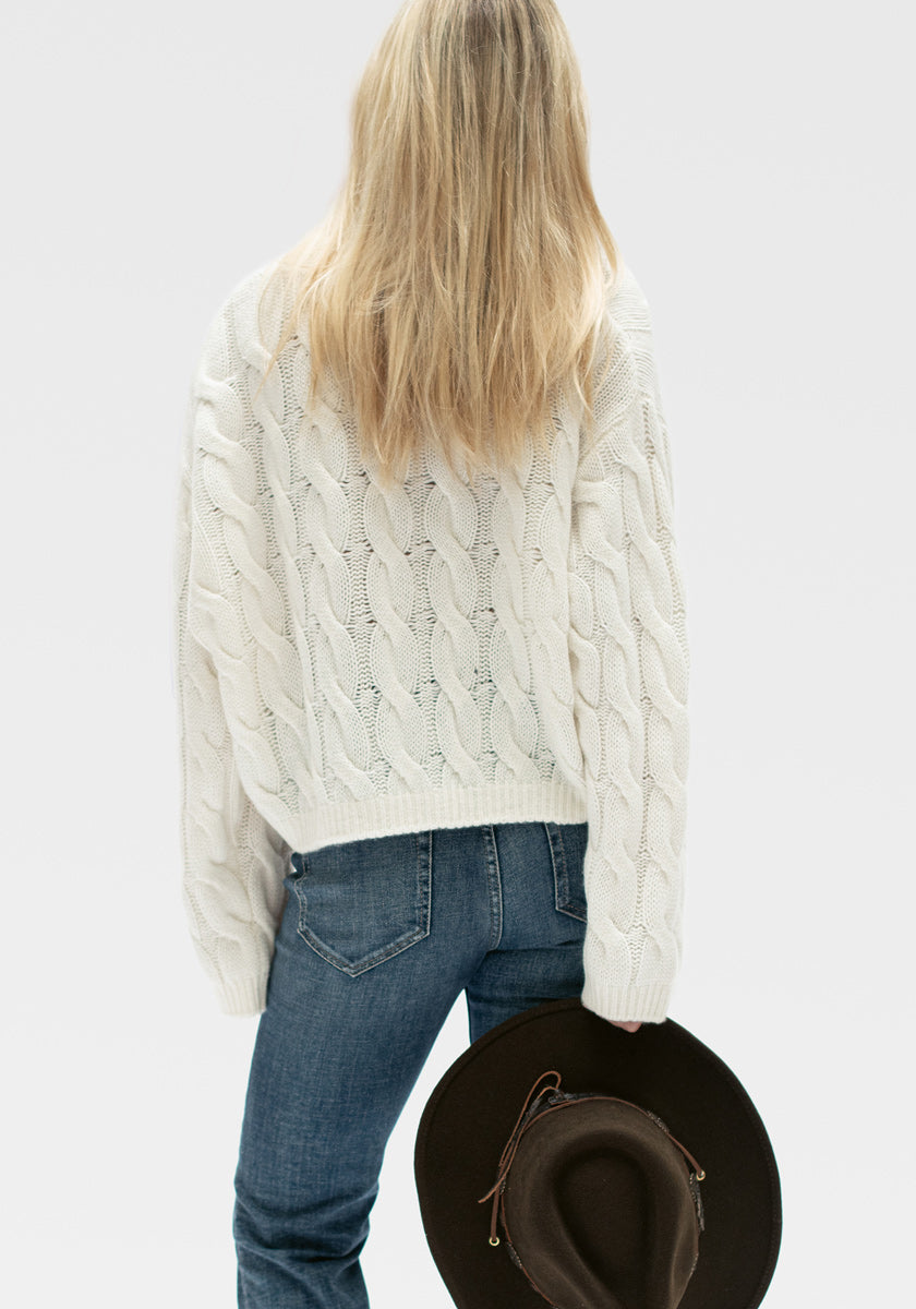 Eine Person mit langen blonden Haaren blickt nach vorne und trägt einen dicken weißen Princess Goes Hollywood-Zopfstrickpullover und blaue Jeans. In ihrer rechten Hand hält sie einen braunen, breitkrempigen Hut vor einem schlichten hellgrauen Hintergrund.