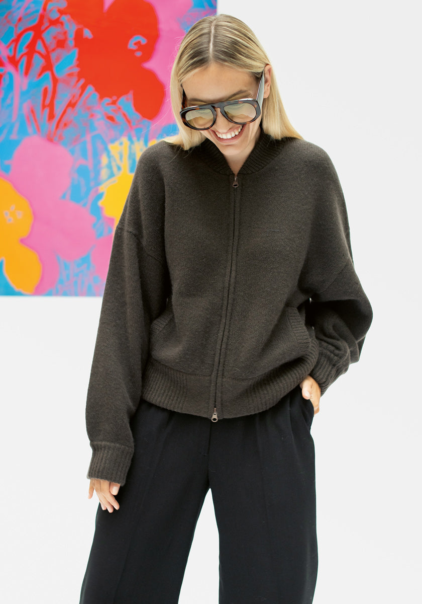 Eine Frau mit glattem blondem Haar trägt eine übergroße Sonnenbrille, eine schwarze Hose und eine dunkle Princess Goes Hollywood Kaschmir Strickjacke mit Stehkragen und zweiseitigem Reißverschluss. Lächelnd, die Hände in den Taschen, steht sie vor einem bunten Blumen-Pop-Art-Gemälde.