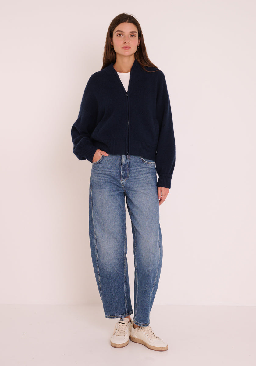 Ein Damenmodel steht vor einem schlichten hellen Hintergrund und trägt eine dunkle marineblaue Princess Goes Hollywood Kaschmir Strickjacke mit Stehkragen über einem weißen Oberteil, eine locker sitzende blaue Jeans und weiße Turnschuhe. Sie hat langes braunes Haar und eine Hand in der Tasche.