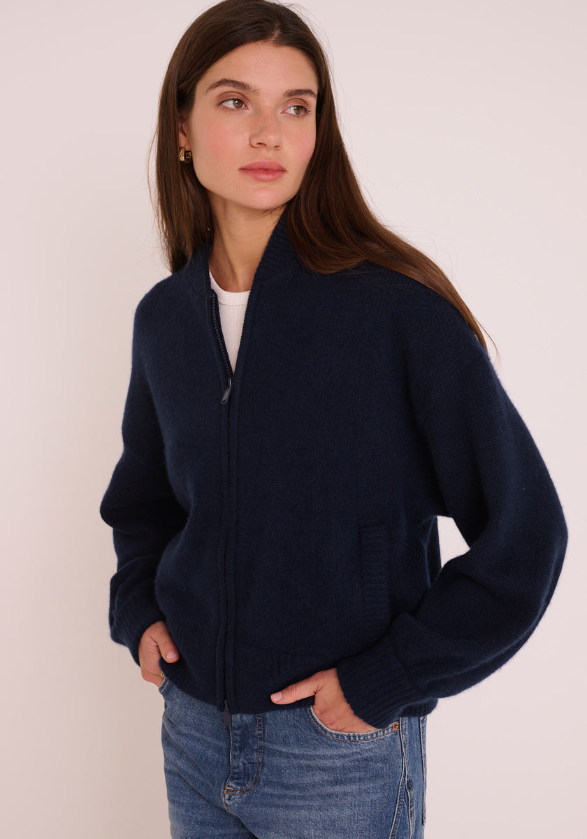 Eine Frau mit langen braunen Haaren trägt eine marineblaue Princess Goes Hollywood Kaschmir Strickjacke mit Stehkragen und zweiseitigem Reißverschluss über einem weißen Oberteil und blauen Jeans. Sie steht vor einem hellen Hintergrund, hat die Hände in den Taschen und einen ruhigen Ausdruck.