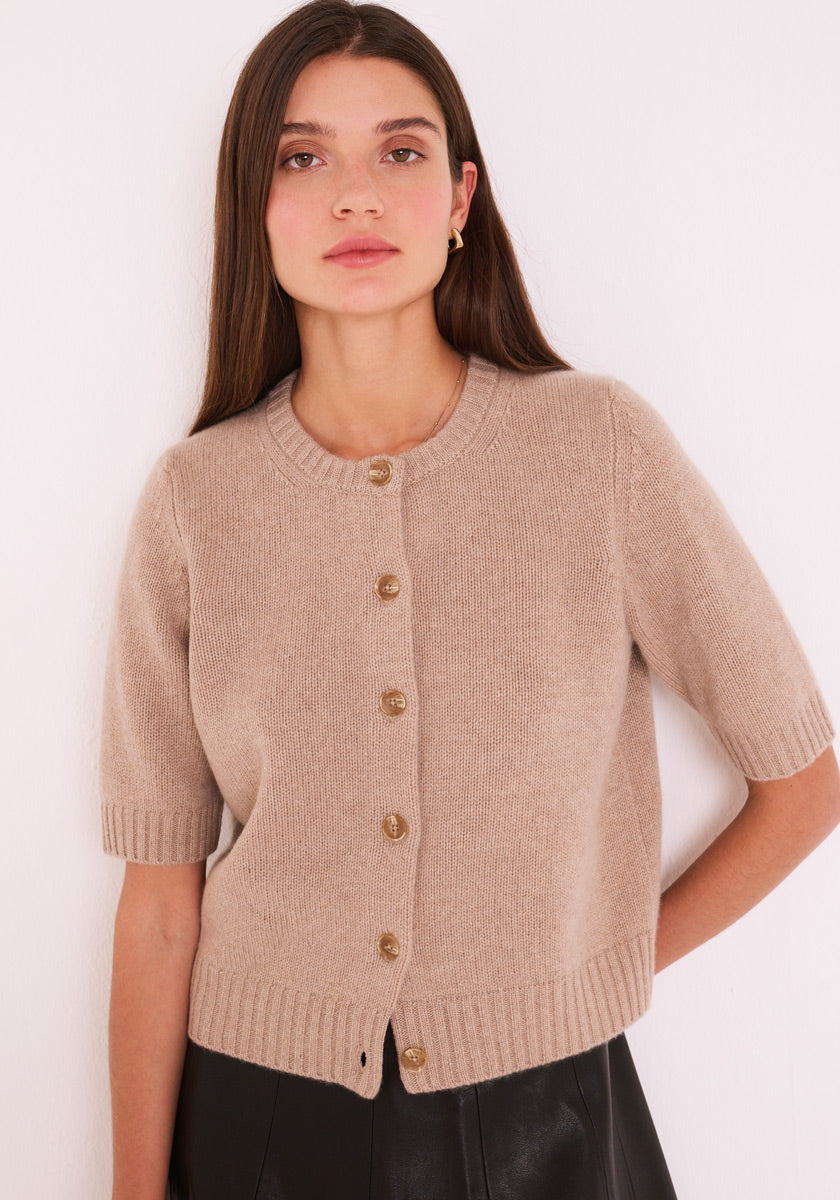 Eine Frau mit langen braunen Haaren trägt einen beigen Princess Goes Hollywood Kaschmir-Strickpullover mit goldenen Knöpfen, gerippten Kanten und Rundhalsausschnitt. Sie steht vor einer weißen Wand und ist mit goldenen Reifen und einem schwarzen Rock oder einer schwarzen Hose ausgestattet.