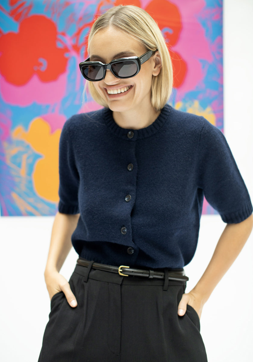 Eine lächelnde Frau mit kinnlangen blonden Haaren und dunkler Sonnenbrille trägt einen marineblauen Princess Goes Hollywood Kaschmir Strickpullover mit kurzen Ärmeln und Rundhalsausschnitt und eine schwarze Hose mit hoher Taille und posiert vor farbenfroher abstrakter Kunst in Rot, Orange, Pink und Blau.