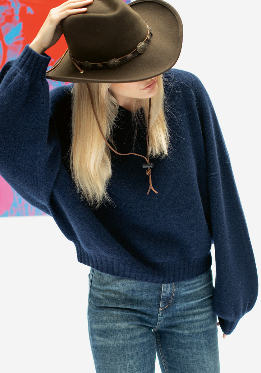 Eine Frau mit langen blonden Haaren trägt einen marineblauen V-Ausschnitt-Pullover von Princess Goes Hollywood, blaue Jeans und einen braunen Cowboyhut. Sie neigt den Hut über ihr Gesicht und steht vor einem bunten abstrakten Kunstwerk im Hintergrund.
