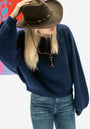 Eine Frau mit langen blonden Haaren trägt einen marineblauen V-Ausschnitt-Pullover von Princess Goes Hollywood, blaue Jeans und einen braunen Cowboyhut. Sie neigt den Hut über ihr Gesicht und steht vor einem bunten abstrakten Kunstwerk im Hintergrund.