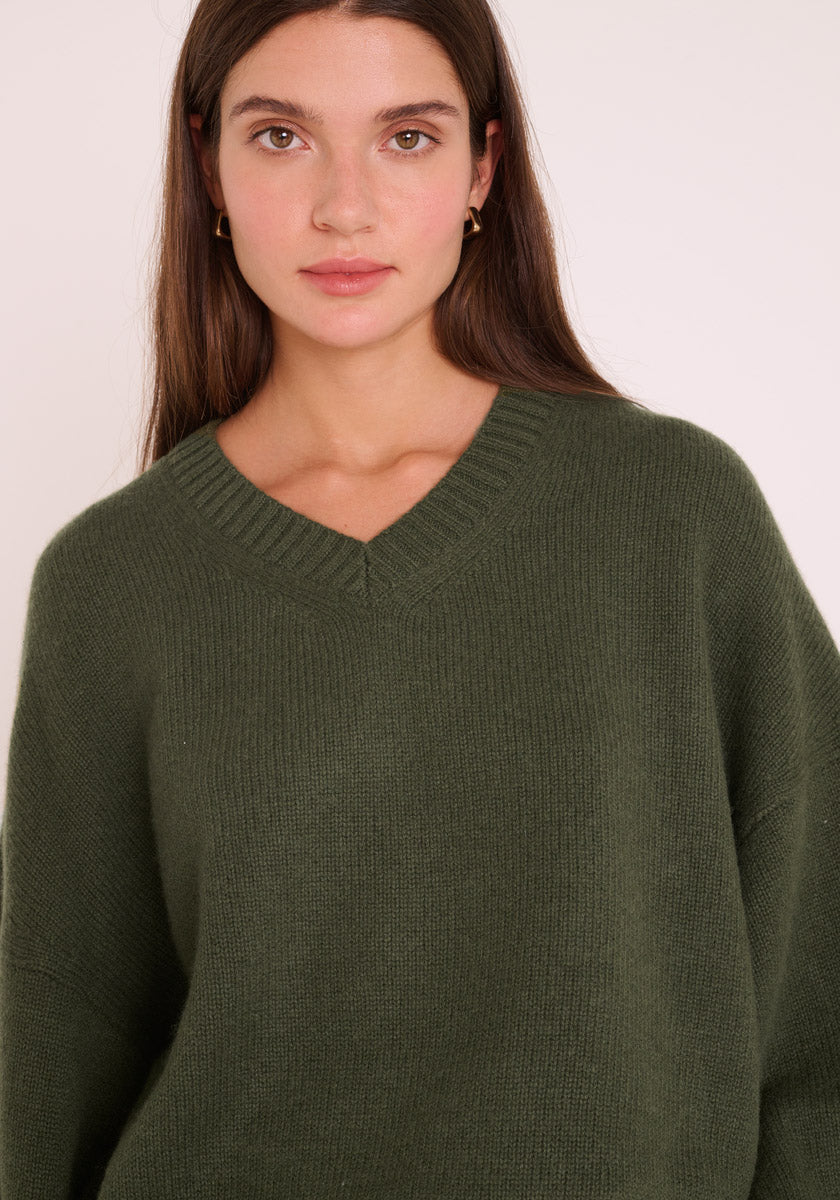 Eine junge Frau mit langen braunen Haaren trägt kleine goldene Ohrringe und einen lockeren, dunkelolivgrünen Kaschmir Strickpullover mit V-Ausschnitt von Princess Goes Hollywood. Sie blickt mit neutralem Gesichtsausdruck vor einem schlichten hellen Hintergrund nach vorne.