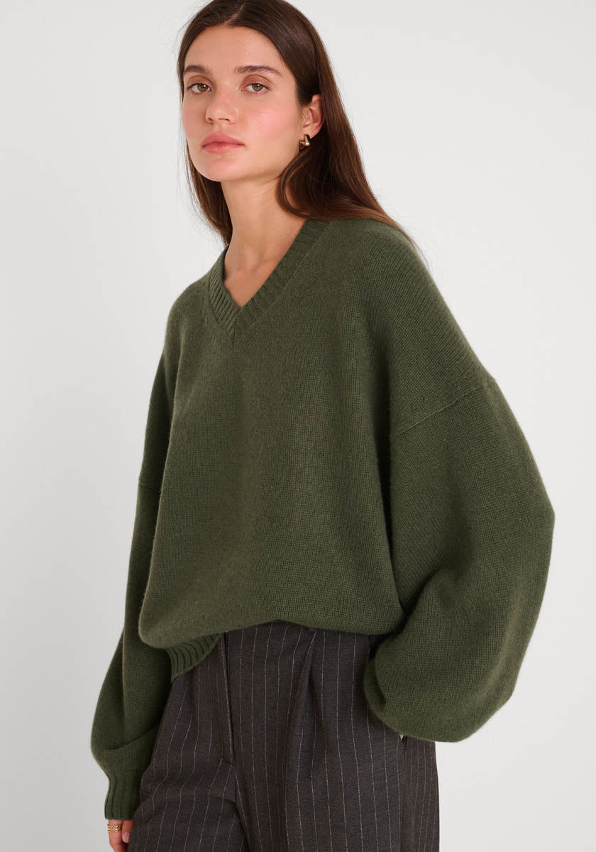 Eine Frau mit glattem braunem Haar trägt einen übergroßen olivgrünen Princess Goes Hollywood Damen Kaschmirpullover mit V-Ausschnitt und langen, weiten Ärmeln, gepaart mit einer anthrazitfarbenen Nadelstreifhose. Sie steht ruhig vor einem schlichten weißen Hintergrund.