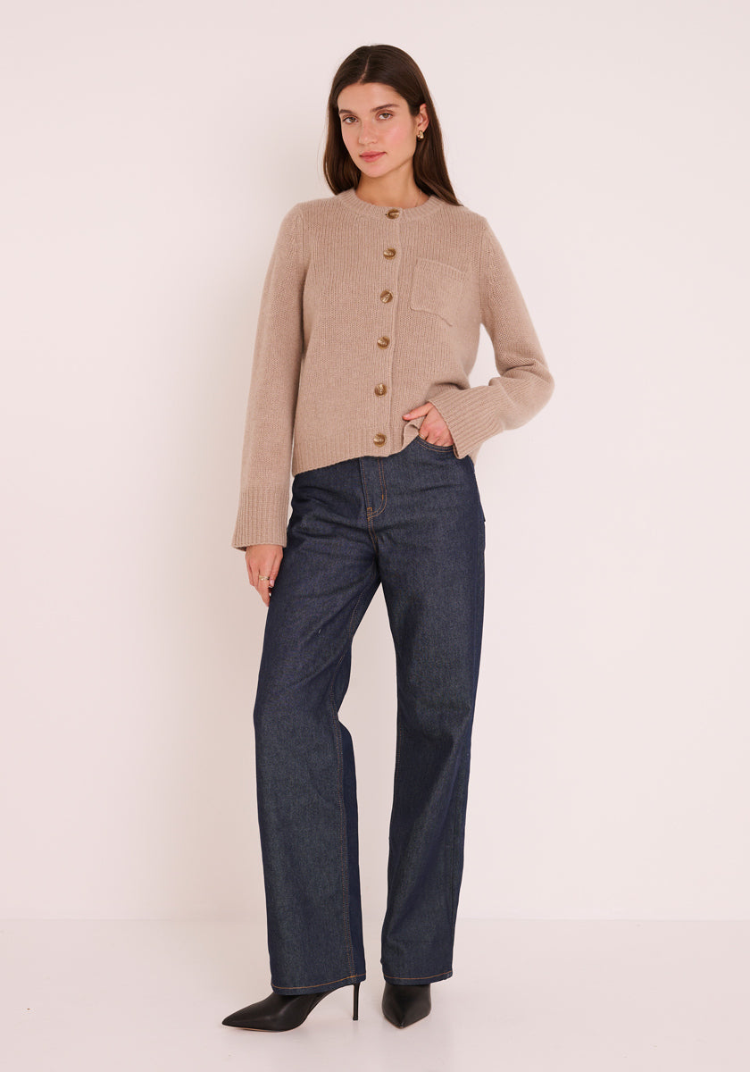 Eine Frau mit glattem braunem Haar und neutralem Gesichtsausdruck trägt eine beigefarbene Princess Goes Hollywood Kaschmir-Strickjacke mit langen Ärmeln, dunkle Jeans mit weitem Bein und schwarze spitze Stiefeletten. Eine Hand steckt in ihrer Tasche, während sie vor einem weißen Hintergrund steht.