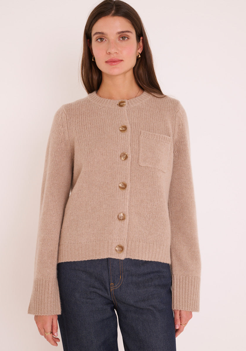 Eine Frau mit glattem braunem Haar trägt eine beigefarbene Princess Goes Hollywood Kaschmir Strickjacke Damen mit Rundhalsausschnitt, Vordertasche und braunen Knöpfen. Dazu trägt sie eine dunkelblaue Jeans vor einem schlichten hellen Hintergrund.