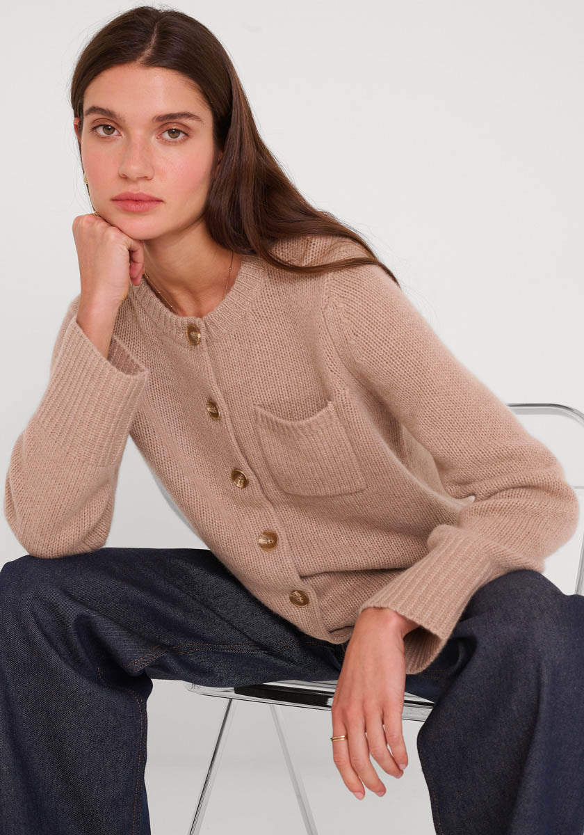 Eine Frau mit langen braunen Haaren sitzt auf einem hellen Stuhl und stützt ihr Kinn auf ihre Hand. Sie trägt eine beigefarbene Princess Goes Hollywood-Kaschmirstrickjacke mit Rundhalsausschnitt und Brusttasche, dazu eine dunkelblaue Jeans mit weitem Bein.