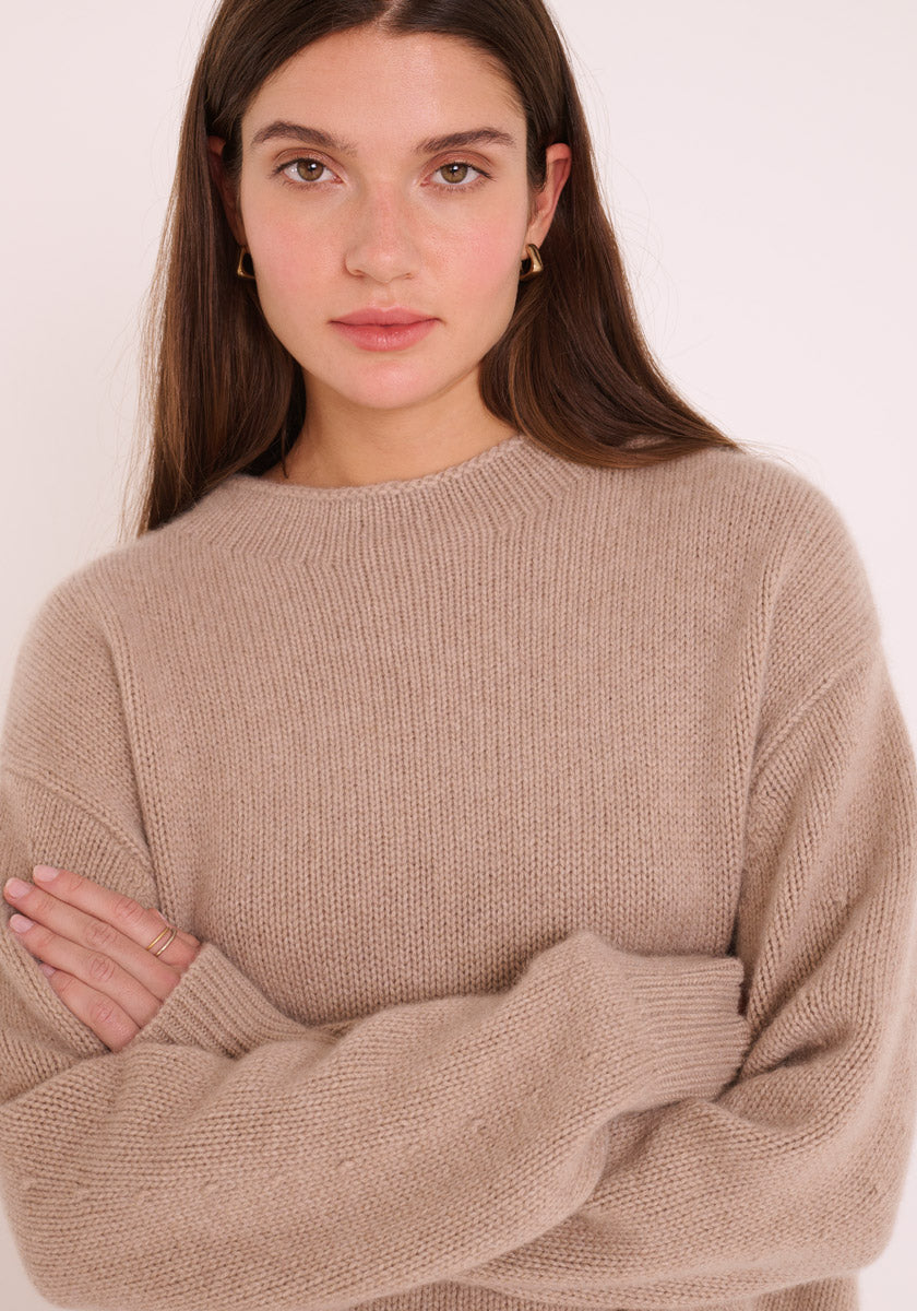 Eine Frau mit langen braunen Haaren, goldenen Ohrringen und neutralem Make-up blickt in die Kamera. Sie trägt einen hellbeigen Damen Pullover mit Stehkragen von Princess Goes Hollywood, die Arme verschränkt, vor einem schlichten hellen Hintergrund.