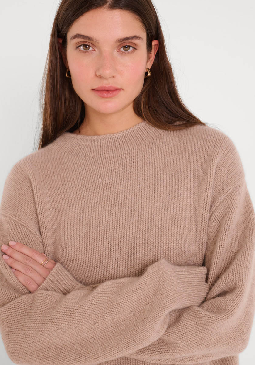 Eine Frau mit langen braunen Haaren trägt einen taupefarbenen Damen Pullover mit Stehkragen von Princess Goes Hollywood. Sie steht mit verschränkten Armen vor einem weißen Hintergrund, trägt kleine goldene Ohrringe und einen neutralen Gesichtsausdruck.