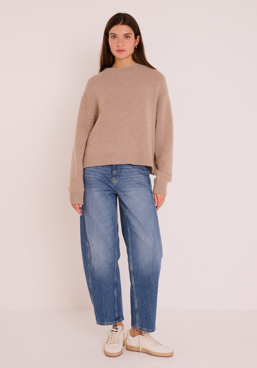 Eine Frau mit langen braunen Haaren steht vor einem schlichten, hellen Hintergrund. Sie trägt einen lässig geschnittenen Princess Goes Hollywood Kaschmir Strickpullover mit Stehkragen, Blue Jeans und weiße Turnschuhe. Ihre Arme sind an den Seiten verschränkt und sie hat einen neutralen Gesichtsausdruck.