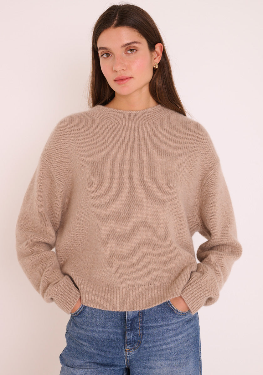 Eine junge Frau mit langen braunen Haaren trägt einen beigen Princess Goes Hollywood Kaschmir Strickpullover Damen Pullover und blaue Jeans. Sie steht vor einem schlichten hellen Hintergrund, die Hände in den Taschen, und schaut ruhig in die Kamera.