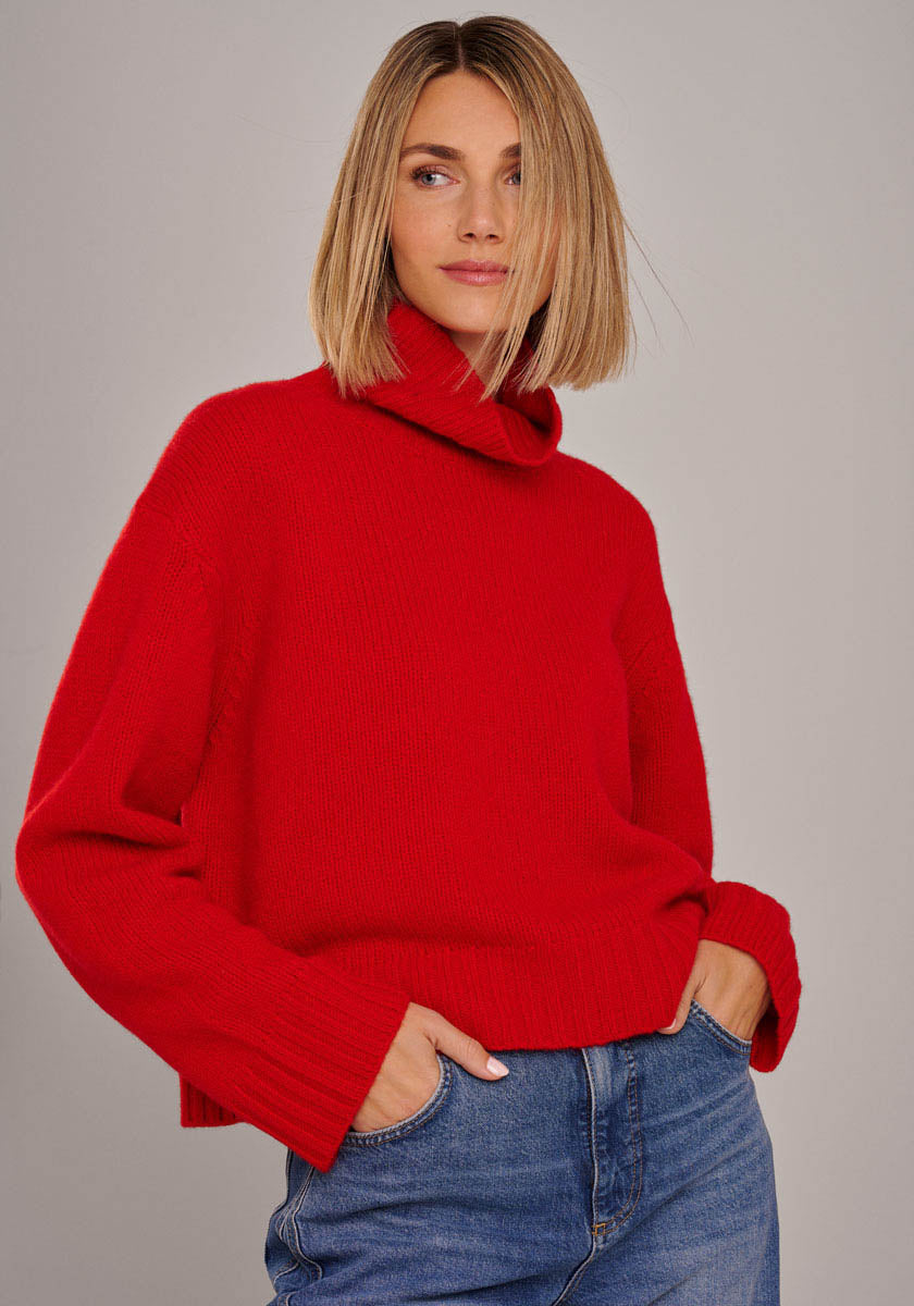 Eine Frau mit glattem, schulterlangem blondem Haar trägt einen knallroten Princess Goes Hollywood Kaschmir Strickpullover mit Rollkragen und langen Ärmeln und Blue Jeans. Sie steht mit den Händen in den Taschen, leicht zur Seite gewandt, vor einem grauen Hintergrund.