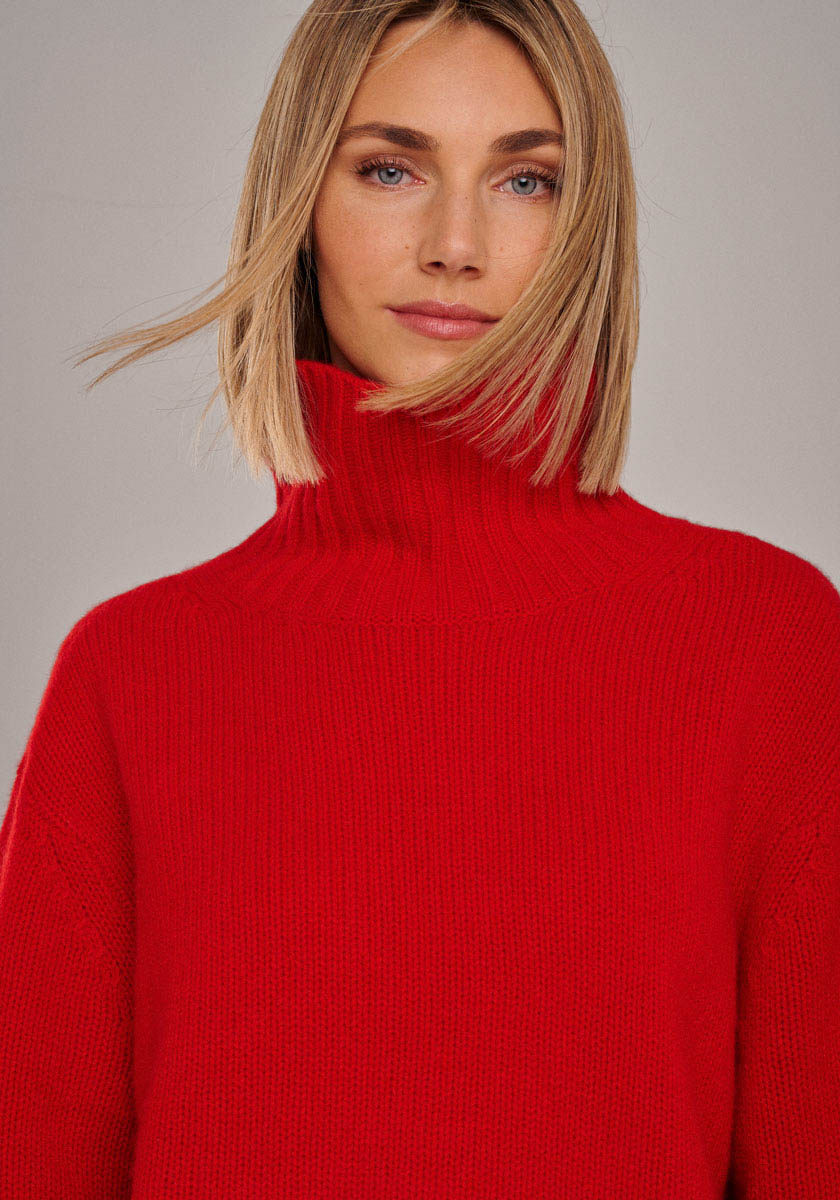 Eine Frau mit glattem, schulterlangem blondem Haar trägt einen leuchtend roten Kaschmir Strickpullover mit Rollkragen und langen Ärmeln von Princess Goes Hollywood. Sie blickt mit einem leichten Lächeln in die Kamera; ihr Haar umrahmt ihr Gesicht sanft vor einem neutralen Hintergrund.