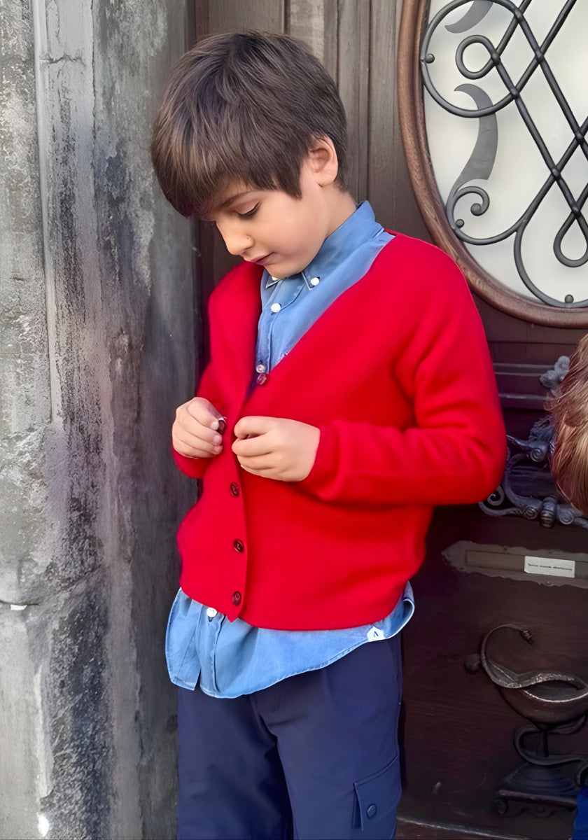 Ein kleiner Junge mit kurzen braunen Haaren knöpft sich eine leuchtend rote Princess Goes Hollywood Strickjacke Kinder mit Rundhalsausschnitt über ein blaues Hemd mit Kragen. Er steht an einer Steinmauer und einer Holztür, sein konzentrierter Ausdruck ist ruhig.