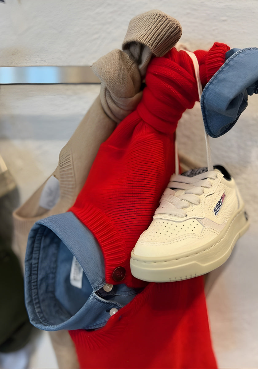 Ein weißer Sneaker mit cremefarbener Sohle hängt an den Schnürsenkeln neben einem roten Pullover, einer beigen Princess Goes Hollywood Kaschmir Strickjacke Kinder und einem blauen Hemd mit Kragen, die alle auf einer strukturierten weißen Wand zusammengeknotet sind.