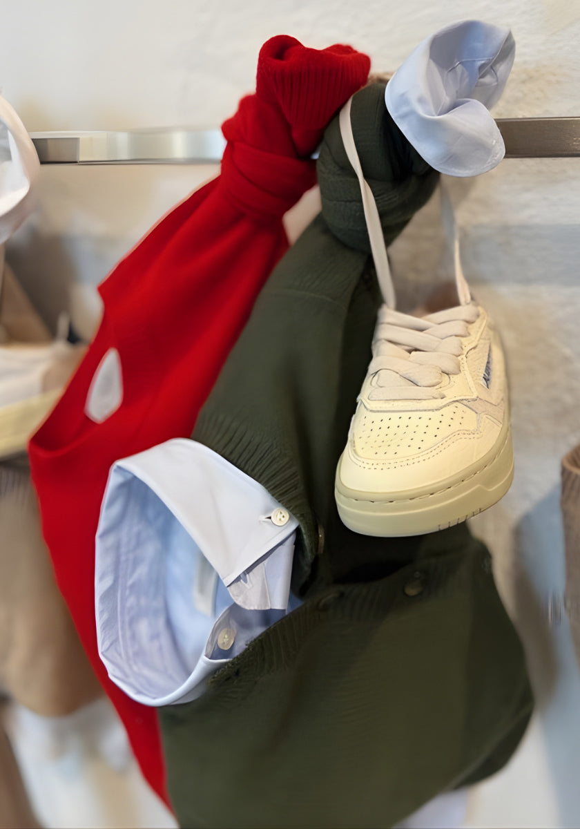 Ein weißer Turnschuh hängt auf einem Regal neben roter und olivgrüner Princess Goes Hollywood Strickjacke Kinder mit Rundhalsausschnitt, Ärmel gebunden. Weiße Hemdmanschetten mit Kragen lugen hervor und zeigen mehrlagige Casual-Styles.