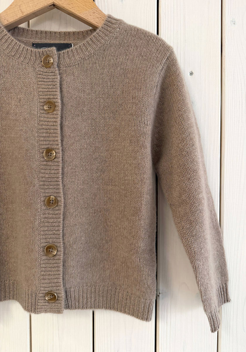 Eine beigefarbene Princess Goes Hollywood Kaschmir Strickjacke Kinder mit langen Ärmeln und einem gerippten Kragen, Manschetten und Saum hängt auf einem Holzbügel vor weißen Platten. Sie hat braune Knöpfe auf der Vorderseite und nur die linke Seite ist abgebildet.