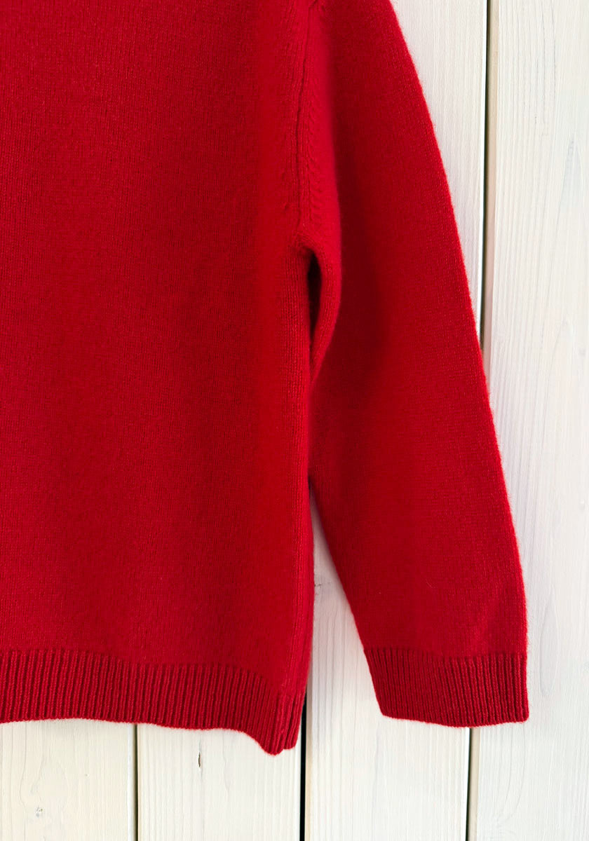 Ein knallroter Princess Goes Hollywood Kaschmir Strickpullover Kinder mit Rundhalsausschnitt hängt an einer hellen Holzwand. Nur der untere Teil und der rechte Ärmel mit Rippbündchen und Saum sind sichtbar und zeigen die weiche, leicht flauschige Textur.