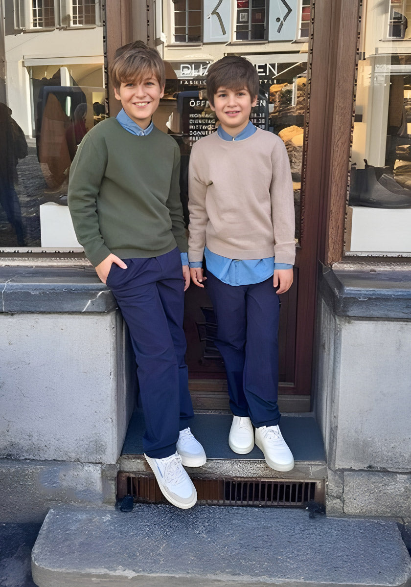 Zwei Jungen stehen vor einem Gebäude in marineblauen Hosen, weißen Turnschuhen und Princess Goes Hollywood Kaschmir Strickpullover Kinder-einer grün, einer beige mit Rundhalsausschnitt. Sie lächeln in die Kamera, im Fenster hinter ihnen spiegelt sich eine Straßenszene.