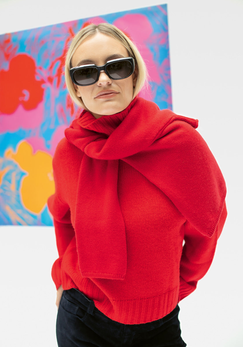 Eine Frau mit schwarzer Sonnenbrille und einem leuchtenden Princess Goes Hollywood Kaschmir Rollkragenpullover, mit einem roten Pullover über den Schultern, steht vor einem abstrakten Gemälde mit kühnen Blumenformen auf blauem Hintergrund.