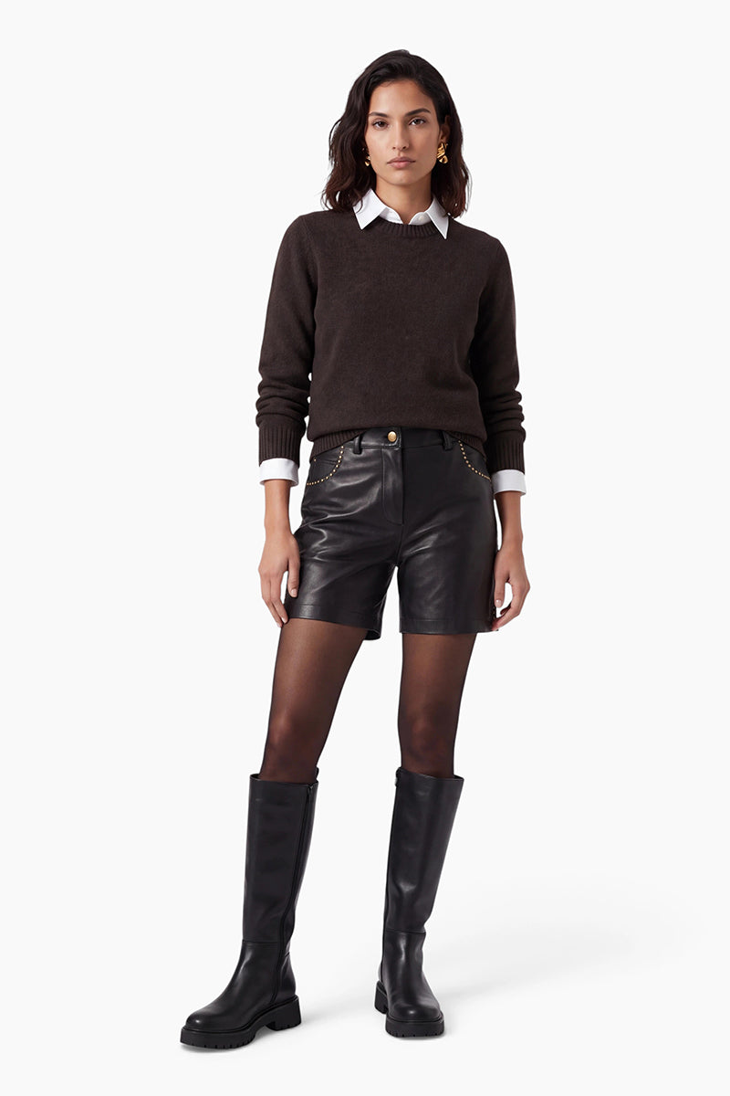 Vor einem schlichten weißen Hintergrund trägt eine Frau einen braunen, langärmeligen Kaschmirpullover über einem weißen Hemd mit Kragen, schwarze Ledershorts mit goldenen Nieten, eine transparente schwarze Strumpfhose und schwarze kniehohe Stiefel.