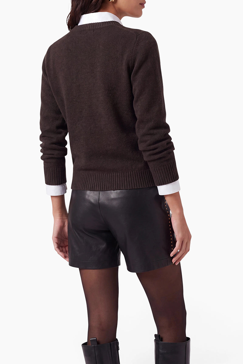 Eine von hinten gezeigte Frau trägt einen dunkelbraunen, langärmeligen Kaschmirpullover über einem weißen Hemd mit Kragen. Dazu trägt sie schwarze Ledershorts, eine transparente schwarze Strumpfhose und schwarze Stiefel vor einem einfarbigen, hellen, cremefarbenen Hintergrund.