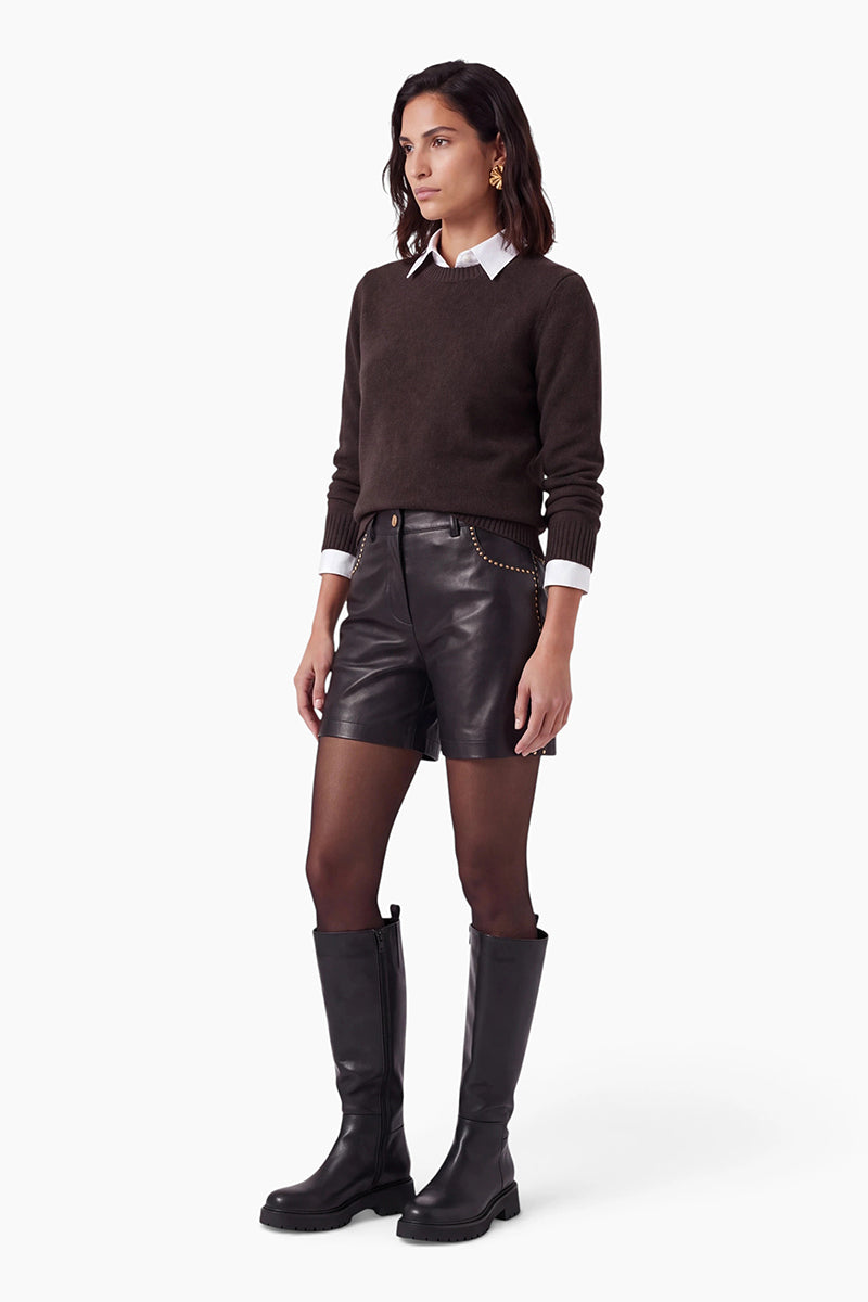 Eine Frau posiert vor einem schlichten weißen Hintergrund in einem dunkelbraunen Kaschmirpullover mit Rundhalsausschnitt, den sie über einem weißen Hemd mit Kragen trägt. Dazu trägt sie schwarze Ledershorts mit goldenen Nieten, schwarze Strumpfhosen und schwarze kniehohe Stiefel.