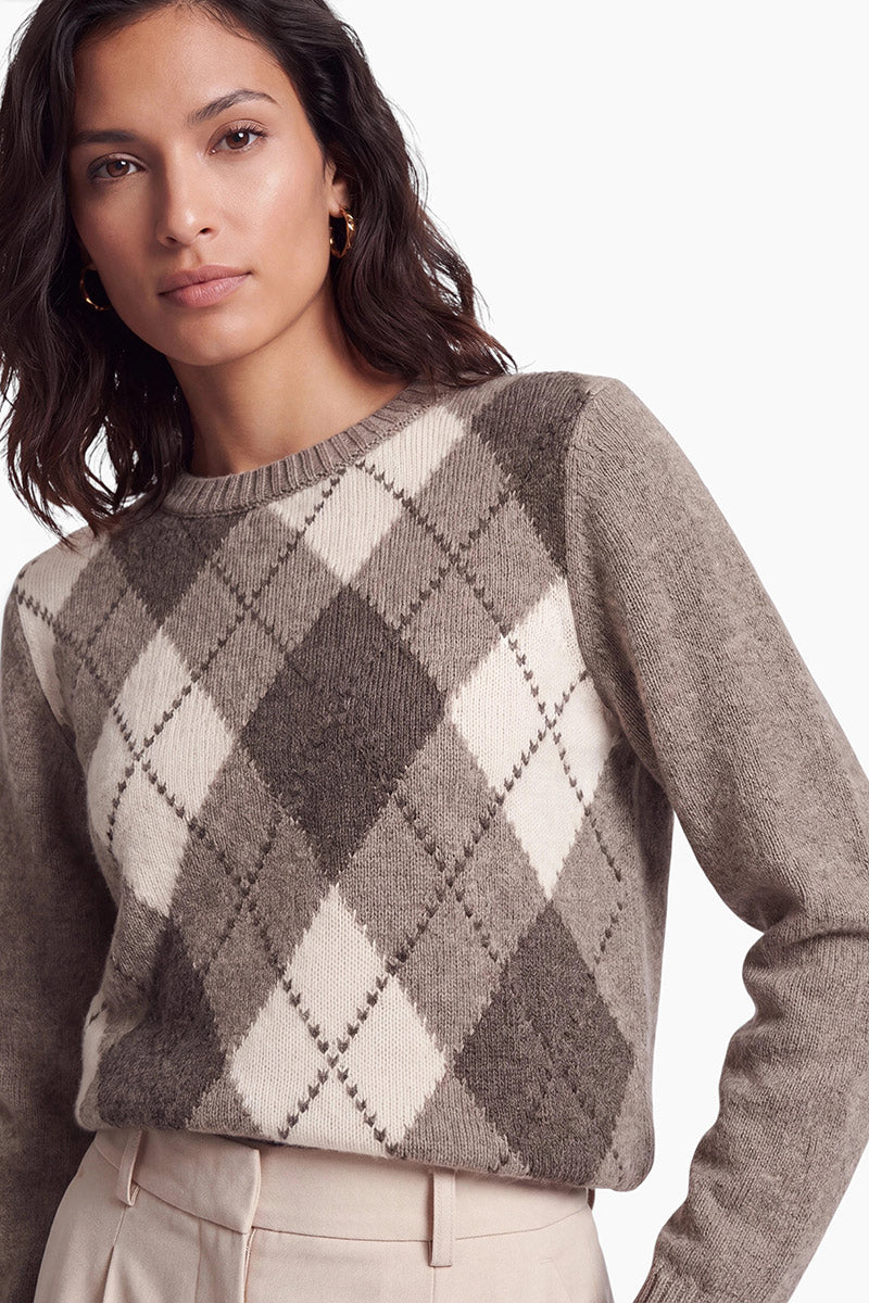 Eine Frau mit dunklem Haar trägt einen braun-weißen Kaschmirpullover mit Argyle-Muster. Vor einem schlichten weißen Hintergrund trägt sie goldene Creolen und eine helle Hose.
