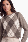 Eine Frau mit dunklem Haar trägt einen braun-weißen Kaschmirpullover mit Argyle-Muster. Vor einem schlichten weißen Hintergrund trägt sie goldene Creolen und eine helle Hose.