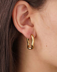Close-up of a person’s ear with straight brown hair tucked behind it. The ear features chunky, irregularly shaped gold or stainless steel earrings from Princess Goes Hollywood, their polished finish catching the light and highlighting smooth skin.