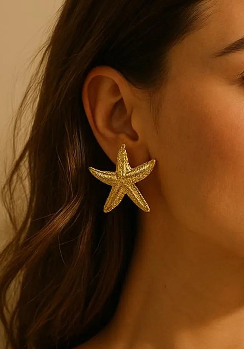 A close-up of a woman’s ear wearing a large, shiny Princess Goes Hollywood Seestern-Ohrstecker with Gelbgold Vergoldung. Her brown hair drapes beside her face, contrasting with her warm skin and the softly lit, neutral background.