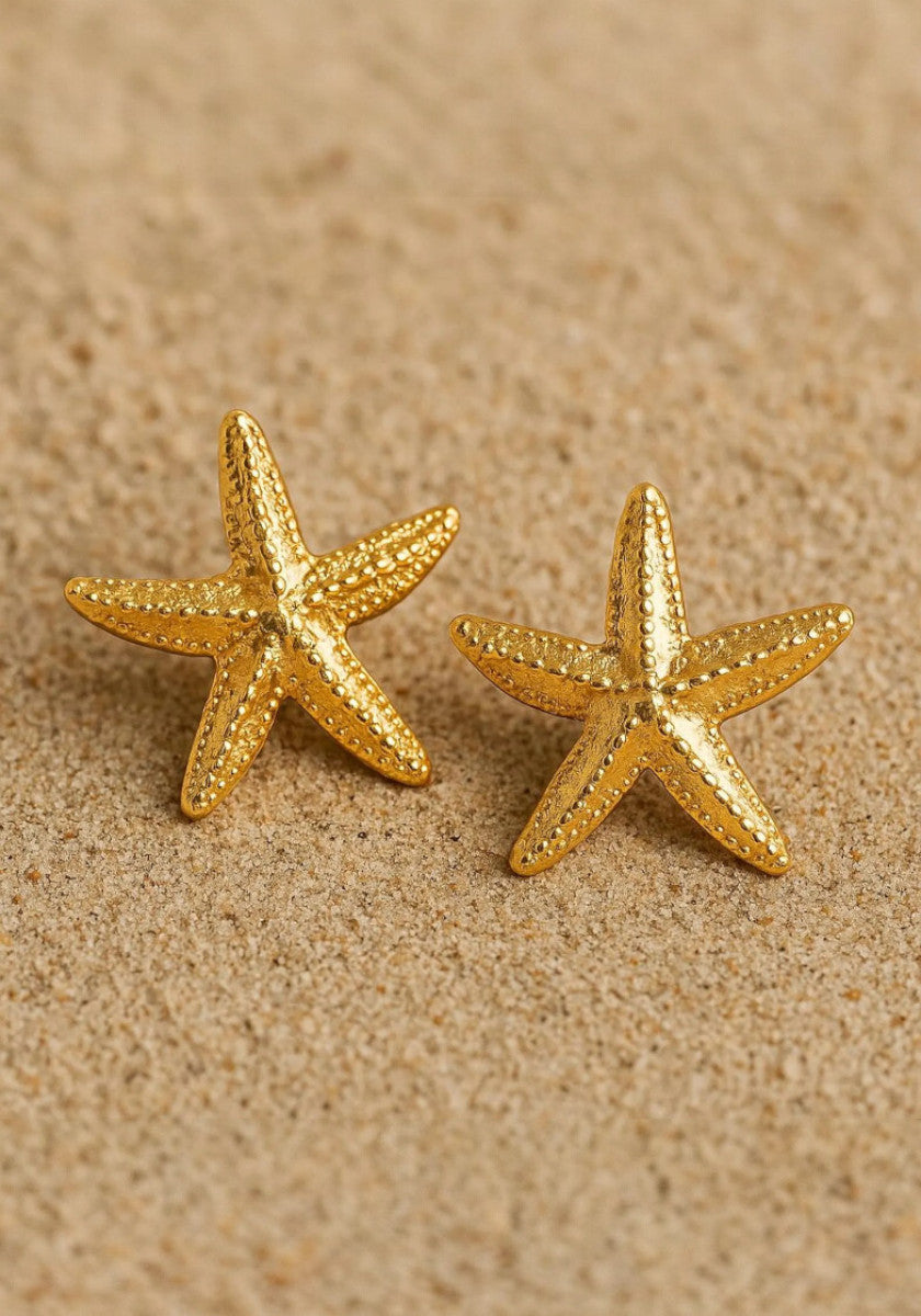 A close-up of two Princess Goes Hollywood Seestern-Ohrstecker aus Edelstahl mit Gelbgold Vergoldung rests on sand, evoking a beach vibe. Placed side by side, they showcase their shiny finish and detailed design.