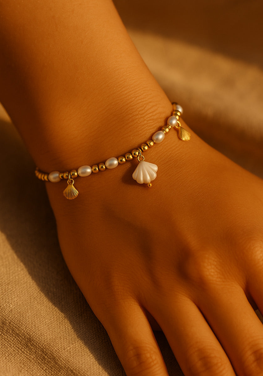 A hand wears a Princess Goes Hollywood Muschel-Armkette with alternating gold and white pearls, three gold shell charms, and a central white shell charm, shown against soft beige fabric in warm lighting that highlights its shine.