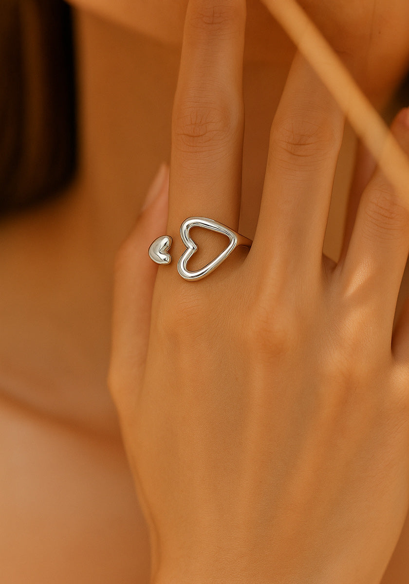 A close-up of a hand wearing a Princess Goes Hollywood Edelstahlring Damen in Herzform: a silver open ring featuring two heart shapes, ideal as a Geschenk mit Herz, set against a softly blurred beige background.