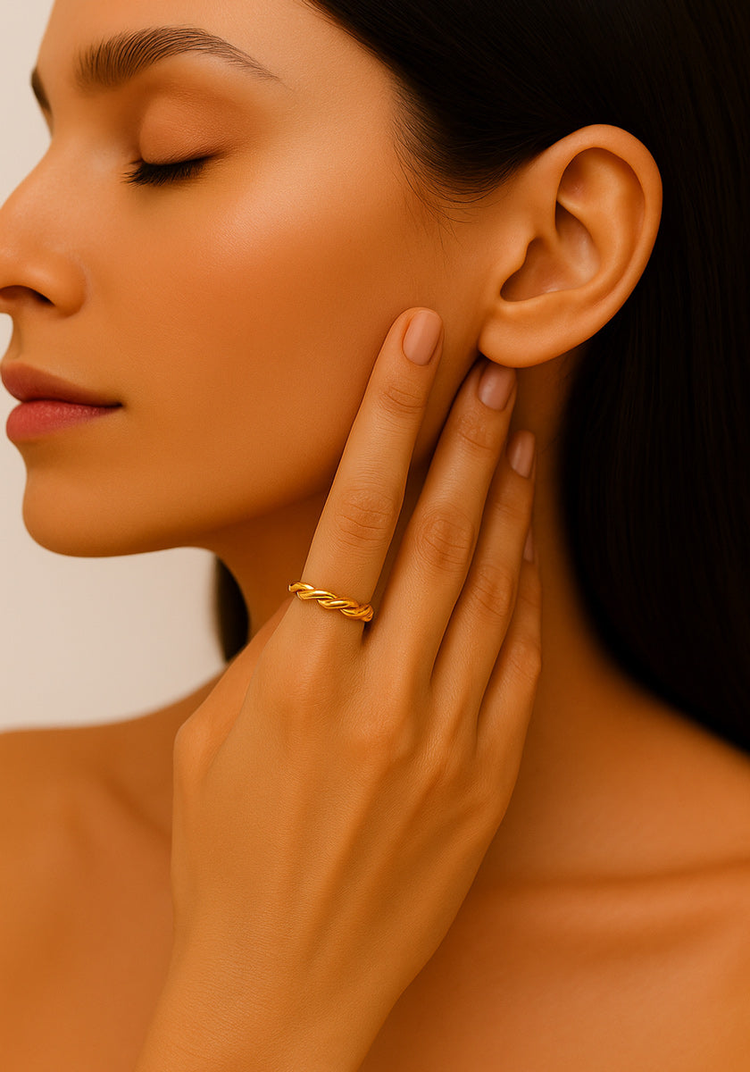 A woman with smooth, tan skin and long dark hair gently touches her face, displaying a Princess Goes Hollywood adjustable stainless steel ring with yellow gold plating. Her closed eyes and natural makeup highlight her serene, elegant features.
