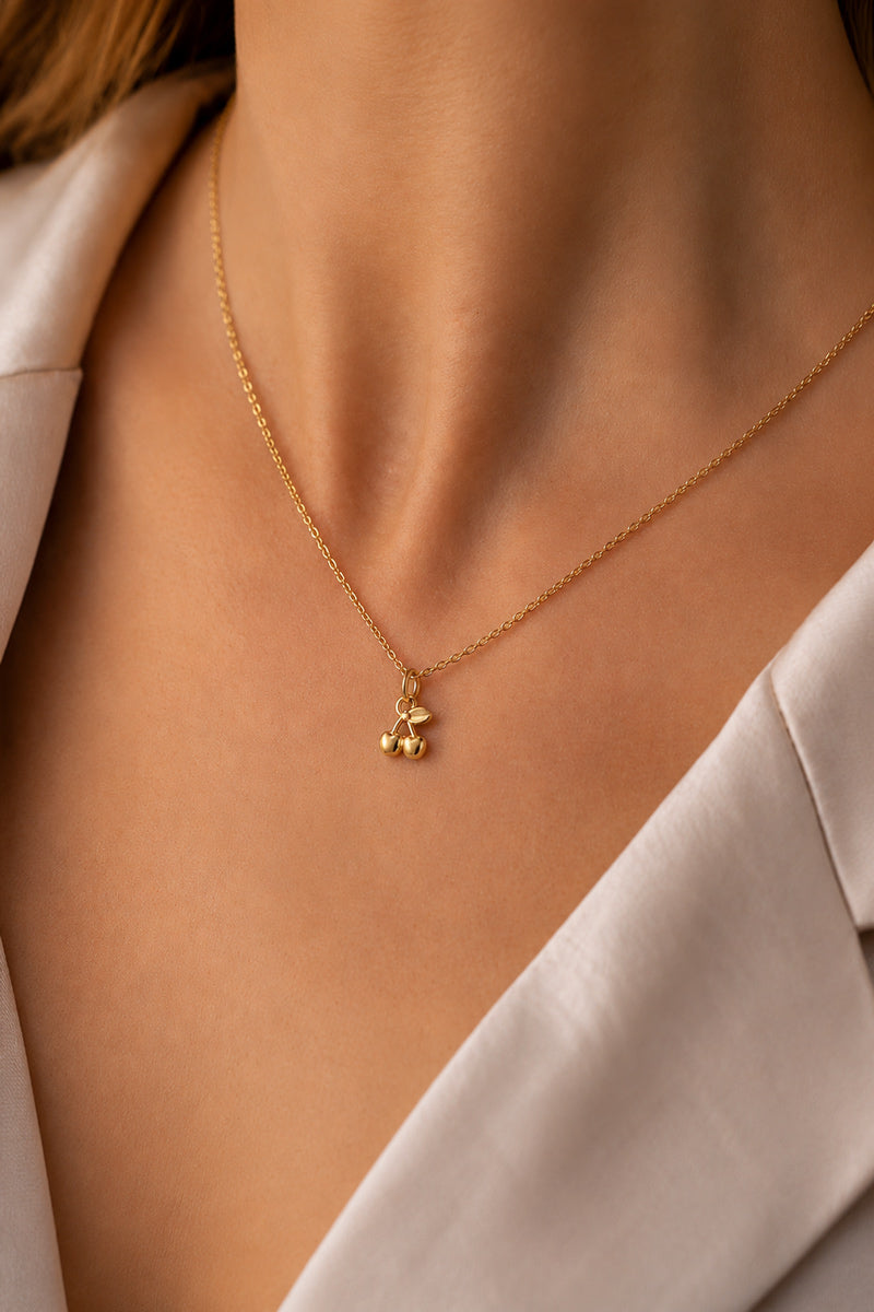 Eine Nahaufnahme zeigt eine Person in einer cremefarbenen Bluse, die den zarten goldenen „Kirsche“-Charm-Anhänger aus Edelstahl von Princess Goes Hollywood trägt; ihre glatte Haut bringt das trendige Accessoire am Ausschnitt besonders gut zur Geltung.