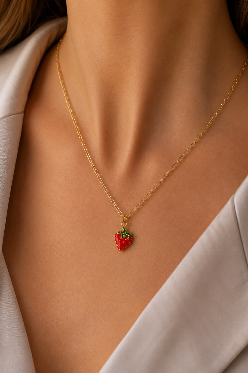 Eine Frau ist von den Schultern abwärts zu sehen; sie trägt einen hellbeigen Blazer und ein weißes Oberteil. Als Accessoire trägt sie eine goldene Kette der Marke „Princess Goes Hollywood“ mit einem kleinen Erdbeer-Anhänger, der mit grünen Blättern verziert ist.