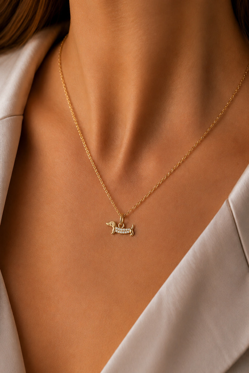 Nahaufnahme des Halses einer Frau, geschmückt mit der goldenen „Dackel Charm“-Halskette von Princess Goes Hollywood, die einen dackelförmigen Edelstahl-Anhänger aufweist. Sie trägt einen hellen Blazer; warmes Licht hebt dieses ideale Geschenk für Hundefans hervor.