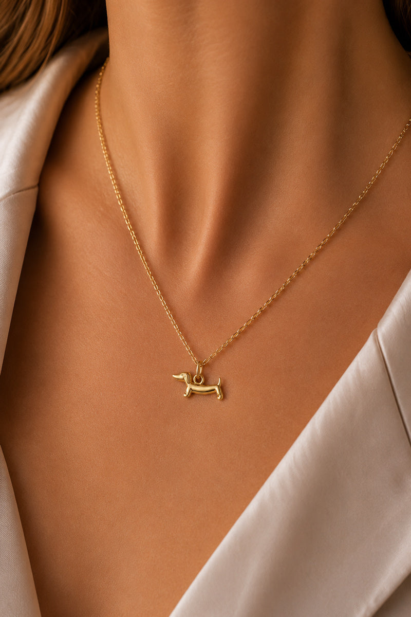 Eine Nahaufnahme einer Frau, die eine zarte Goldkette mit einem Dackel-Anhänger von Princess Goes Hollywood trägt. Sie trägt eine cremefarbene, satinartige Bluse mit einem weichen Kragen vor einem sanft unscharfen Hintergrund. Schmuck für Tierliebhaber.