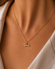 Eine Nahaufnahme einer Frau, die eine zarte Goldkette mit einem Dackel-Anhänger von Princess Goes Hollywood trägt. Sie trägt eine cremefarbene, satinartige Bluse mit einem weichen Kragen vor einem sanft unscharfen Hintergrund. Schmuck für Tierliebhaber.