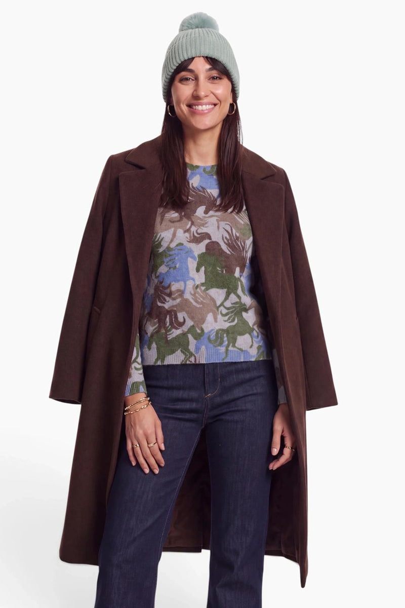 Eine lächelnde Frau trägt eine salbeigrüne Mütze aus Rippstrick, einen dunkelbraunen Mantel und einen blau-braunen Pullover mit Pferdemuster. Vor einem schlichten weißen Hintergrund trägt sie eine dunkelblaue Jeans und goldenen Schmuck.