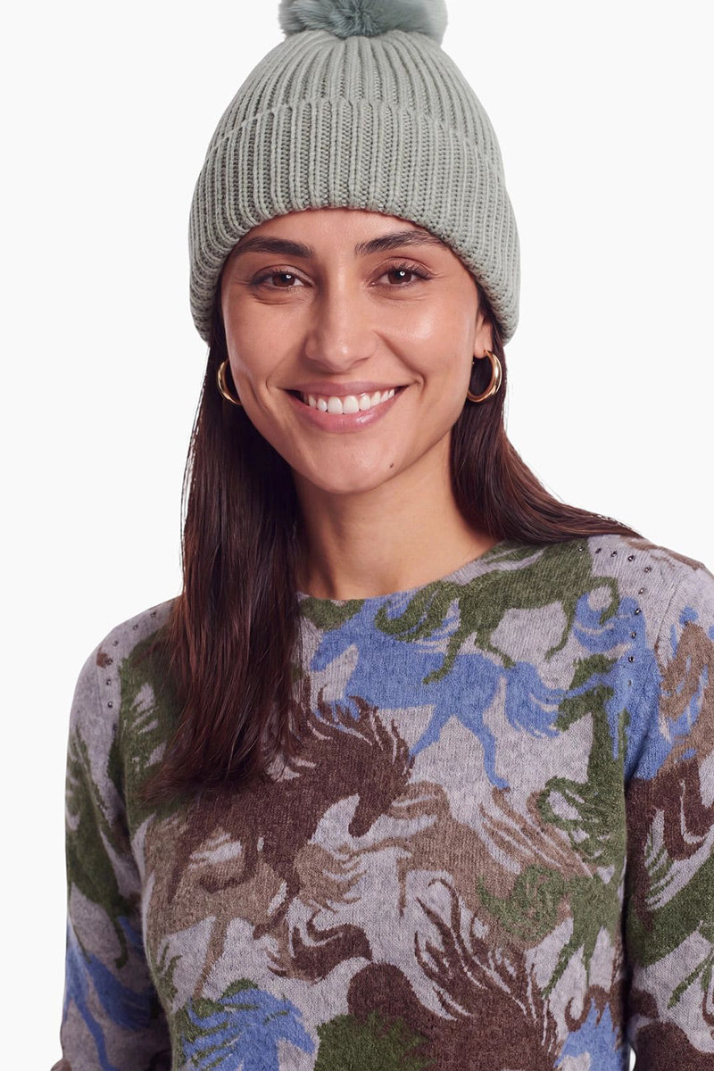 Eine lächelnde Frau trägt eine salbeigrüne Rippstrick-Mütze mit einem Bommel. Sie hat langes braunes Haar, trägt goldene Creolen und einen grauen Pullover mit einem Muster aus Pferden in Braun-, Blau- und Grüntönen.
