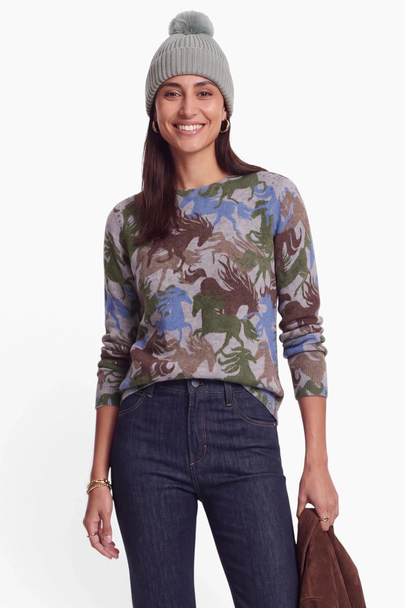 Eine lächelnde Frau trägt eine graue, gerippte Mütze mit Bommel und einen langärmeligen Pullover mit einem Camouflage-Muster mit Pferdemotiv. Sie trägt eine dunkelblaue Jeans vor einem schlichten weißen Hintergrund.