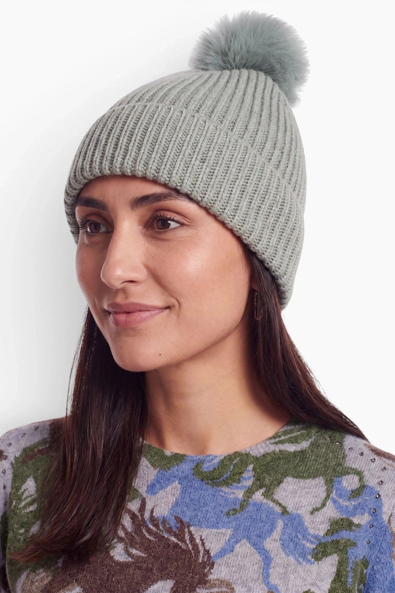 Eine Frau mit langen, dunklen Haaren trägt eine salbeigrüne, gerippte Strickmütze mit einem passenden Bommel aus Kunstfell. Sie ist leicht zur Seite gedreht und trägt einen mehrfarbig gemusterten Pullover vor einem schlichten weißen Hintergrund.